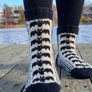 Puede incluir: Un par de calcetines de punto con un patrón en blanco y negro. Los calcetines tienen puños, talones y punteras negros. El patrón principal es un diseño repetitivo de formas negras sobre un fondo blanco. Los calcetines se llevan sobre una superficie de madera.