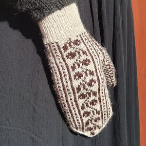 Könnte beinhalten: Ein gestrickter Fäustling mit einem braunen und cremefarbenen Muster. Das Bündchen ist cremefarben und der Fäustling hat ein sich wiederholendes Blumendesign in Braun. Der Fäustling wird an einer Hand getragen, wobei ein dunkelgrauer Ärmel sichtbar ist.