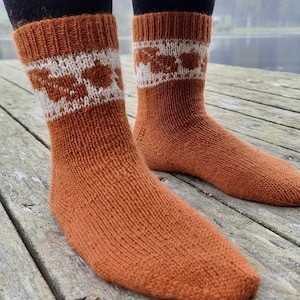 May include: A pair of knitted socks in a warm orange-brown colour. The socks have a white and brown geometric pattern around the cuff. The socks are ribbed and appear to be made of a soft, warm material, ideal for cold weather.