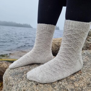 May include: A pair of light gray knitted socks with a diamond pattern. The socks are worn with black leggings and are resting on a large rock near a body of water. The socks are made of a soft, textured yarn.