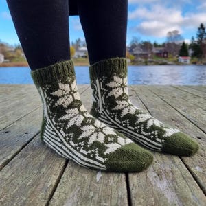 May include: A pair of olive green and white knit socks with a snowflake pattern. The socks have a ribbed cuff and toe, and are worn over black leggings. The socks are on a wooden dock with a lake in the background.