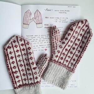 Pode incluir: Um par de luvas de malha feitas à mão em creme e bordô. As luvas apresentam um padrão floral repetido e punhos canelados. Um caderno com anotações manuscritas e um esboço das luvas é visível.
