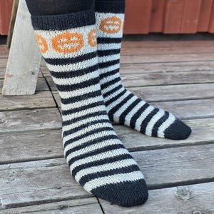 May include: A pair of mid-calf knit socks featuring a black and white horizontal striped pattern throughout the foot and leg. The upper cuff area includes a band of orange jack-o'-lantern pumpkin motifs. The toe and top cuff are solid black.
