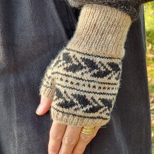 Op de afbeelding: Beige en zwarte gebreide vingerloze handschoenen met een geometrisch patroon. De handschoenen hebben een geribbelde manchet en zijn gemaakt van een zacht, getextureerd garen. Het ontwerp heeft zwarte ruitvormen en kleine driehoeken.