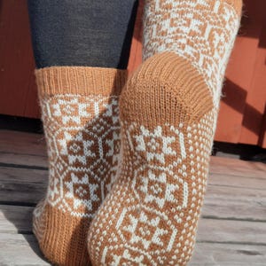 May include: A pair of knitted socks in shades of brown and white. The socks feature a geometric pattern and are visible from the ankles down. The socks are made of a soft, warm material, perfect for cold weather.