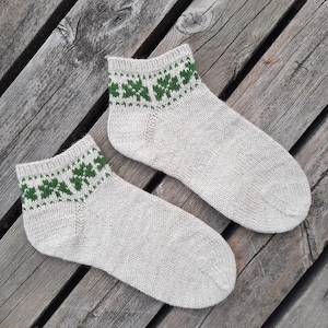 May include: A pair of light gray knitted socks with green geometric patterns on the cuffs. The socks are displayed on a weathered wooden surface, showcasing a handmade, cozy aesthetic.