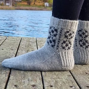 May include: A pair of gray knitted socks with a geometric black pattern. The socks are made of a soft, textured yarn and have a ribbed cuff. The socks are shown on a wooden dock with a lake in the background.