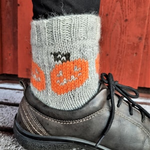 May include: Gray knitted socks with orange pumpkin designs are visible, worn with a dark brown leather shoe. The socks are pulled up over the ankle, with the pumpkin design clearly visible. The shoe has black laces.