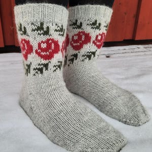 May include: A pair of light gray knitted socks with a floral pattern. The socks feature red roses and green leaves, with a ribbed cuff and a textured knit design. The socks are shown on a snowy surface.