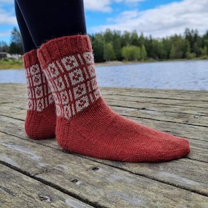 May include: A pair of red knitted socks with a geometric white pattern on the cuffs. The socks are made of a thick, warm material, perfect for cold weather. The socks are shown on a wooden dock with a lake and trees in the background.