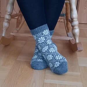 May include: A pair of gray and white patterned socks. The socks feature a repeating white floral design on a gray background. The socks are worn with dark blue leggings. The image is taken indoors, with a wooden rocking chair in the background.