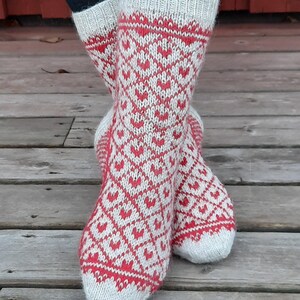 May include: A pair of knitted socks with a red and white heart pattern. The socks have a white cuff and toe, and the heart design covers the rest of the sock. The socks are on a wooden surface.