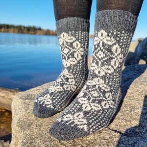 May include: A pair of gray and white patterned socks. The socks feature a repeating floral design in white against a gray background. The socks are worn with dark gray leggings and are resting on a rock near a body of water.