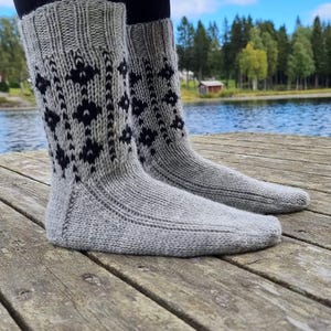 May include: A pair of gray knitted socks with a black diamond pattern. The socks are thick and appear warm, with a ribbed cuff and a patterned design on the front. The socks are on a wooden dock.