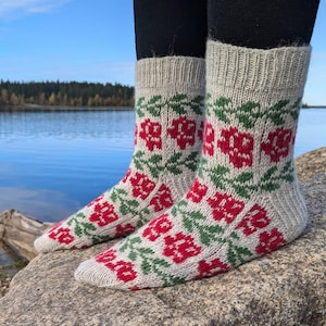 May include: A pair of knitted socks with a floral pattern. The socks are light gray with red flowers and green leaves. The socks are knee-high and are displayed outdoors.