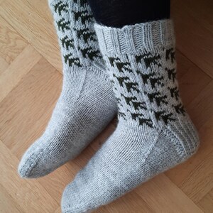 May include: A pair of light gray knitted socks with a dark green geometric pattern on the cuff. The socks are made of a soft, textured material and are designed to be worn on the feet. The socks are on a wooden surface.