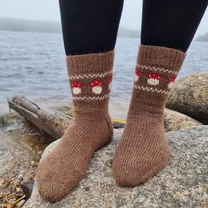 May include: Brown knit socks with a repeating pattern of red and white mushrooms. The socks are worn with black leggings. The socks are displayed on a gray rock near a body of water.