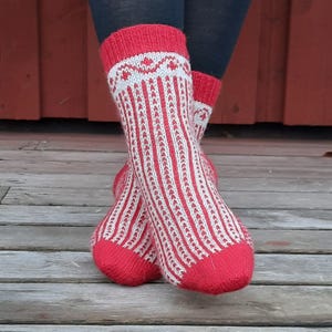 May include: A pair of red and white knitted socks. The socks have a red cuff with a white geometric pattern. The body of the socks features vertical white stripes against a red background. The socks are displayed on a wooden surface.