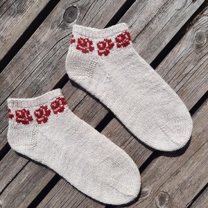 May include: A pair of light gray ankle socks with a red floral pattern at the cuff. The socks are made of a knitted material and are displayed on a weathered wooden surface. The socks are a cozy accessory.