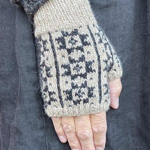 Könnte beinhalten: Ein Paar fingerlose Handschuhe mit beigefarbener Basis und schwarzem geometrischem Muster. Die Handschuhe haben ein geripptes Bündchen am Handgelenk und eine Daumenöffnung aus strukturiertem Strickmaterial, das den Handrücken bedeckt.