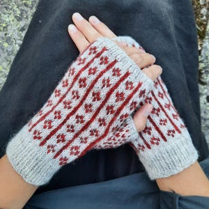 Könnte beinhalten: Ein Paar gestrickte Fäustlinge in Hellgrau mit einem roten Blumenmuster. Die Handschuhe haben vertikale rote Streifen mit kleinen roten Blumenmustern. Die Bündchen sind einfarbig hellgrau.
