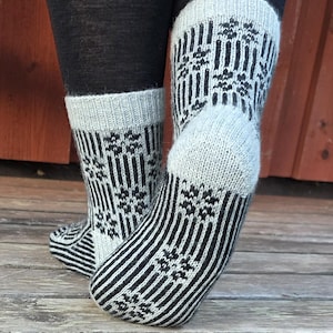 May include: A pair of mid-calf knit socks featuring a repeating pattern of black vertical stripes and small black floral motifs on a light gray background. The socks have a solid light gray heel and a ribbed cuff at the top edge.