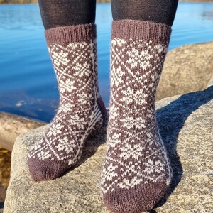 May include: A pair of mid-calf knit socks featuring a brown and white geometric floral pattern. The socks have a ribbed cuff at the top and a solid brown toe section, crafted with a visible wool-like texture throughout the knit fabric.