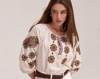 May include: A white embroidered blouse with a geometric pattern in red, orange, and green. The blouse has a round neckline and three-quarter sleeves. The model is wearing a brown braided belt and white denim shorts.