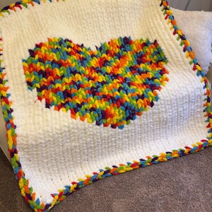 May include: A white, plush blanket with a large, colorful heart design in the center. The heart is made of vibrant rainbow-colored yarn. The blanket has a rainbow-colored border, adding a cheerful touch. This handmade blanket is perfect for snuggling.