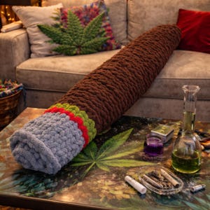 Puede incluir: Un gran cojín marrón tejido a ganchillo con forma de porro de cannabis descansa sobre una mesa con un diseño de hoja de cannabis. El cojín tiene detalles grises, rojos y verdes. Un bong de vidrio y otros accesorios también están sobre la mesa.