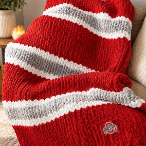 May include: A chunky knit throw blanket featuring thick red, white, and gray horizontal stripes. The blanket is crafted from soft, plush yarn and includes a small, embroidered Ohio State logo patch positioned in the lower right corner of the fabric.