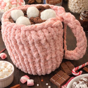 May include: A large, pink, knitted mug filled with hot chocolate and topped with white marshmallows. The mug is surrounded by ingredients like chocolate bars, marshmallows, and a can of Swiss Miss hot chocolate, creating a cozy, inviting scene.