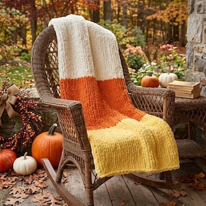 May include: A chunky knit blanket in the colors of candy corn, draped over a wicker rocking chair. The blanket features white, orange, and yellow stripes. Pumpkins and fall foliage surround the chair, creating a cozy autumn scene.