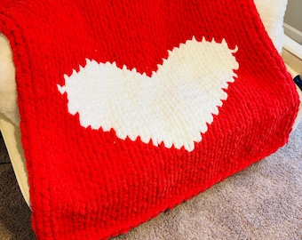 May include: A red knitted throw blanket with a white heart design. The blanket is made of chunky yarn and has a soft, fuzzy texture.
