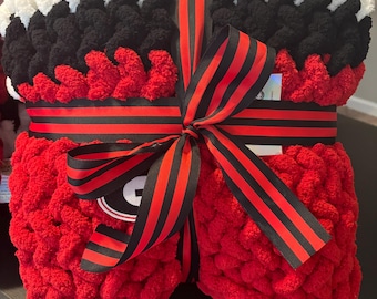 May include: A hand-knitted chunky yarn blanket featuring horizontal stripes of red, black, and white. The blanket is folded and secured with a black and red striped ribbon tied in a bow, with a small circular logo tag visible near the center.