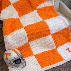 May include: A handmade, chunky knit blanket in a checkerboard pattern of orange and white. A football helmet with an orange "T" logo rests on the blanket. The blanket is displayed on a white chair.