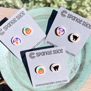May include: Three cards with the text "Sparkle Slice" and a logo of a lemon slice. Each card has two small, round, white earrings with different Halloween-themed designs: a pumpkin, a ghost, a black cat.