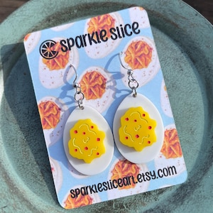 May include: A pair of dangle earrings featuring a white oval with a yellow egg yolk and red pepper flakes. The earrings are on a card with the text "sparkle slice" and "sparklesliceart.etsy.com".