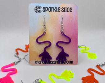May include: Purple acrylic earrings shaped like a hand with a wavy line leading to the hand. The earrings are on a card with the text "sparkle slice" and a website address.