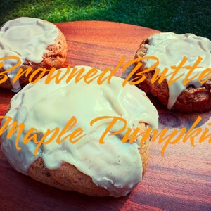 May include: Three baked goods with white frosting on a wooden surface. The text "Browned Butter Maple Pumpkin" is written in orange script across the image. The baked goods are round and appear to be muffins or scones.