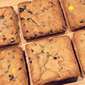 Op de afbeelding: Zes vierkante chocoladekoekjes met kleurrijke snoepjes op een houten oppervlak. De koekjes zijn goudbruin en hebben een licht knapperige textuur.