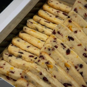 May include: A box of shortbread cookies, arranged in neat rows. The cookies are rectangular with rounded edges and studded with dried cranberries and orange zest. The cookies have a golden-brown color.