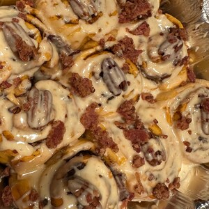 Könnte beinhalten: Nahaufnahme einer Pfanne mit Kürbis-Zimtschnecken, die mit cremigem weißem Zuckerguss, gehackten Pekannüssen und knusprigen Speckwürfeln belegt sind.