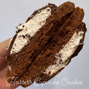 Puede incluir: Una galleta Hostess Cupcake Cookie cortada por la mitad, revelando una galleta de chocolate con un relleno de glaseado blanco. La galleta está cubierta de chocolate y chispas.