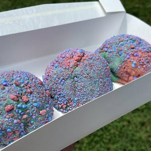 Puede incluir: Tres dulces redondos y coloridos en una caja blanca. Cada dulce está cubierto con una mezcla de chispas azules, rosas y verdes. Los dulces son un postre vibrante y llamativo.