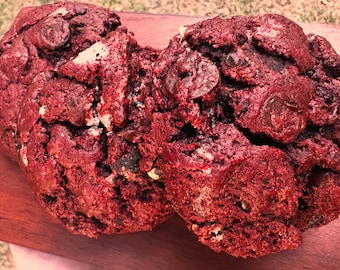 May include: Two large red velvet cookies with chocolate chips and white chocolate chunks. The cookies are on a wooden surface.