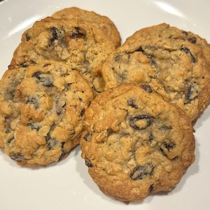 Puede incluir: Primer plano de cinco galletas de avena y pasas en un plato blanco. Las galletas son de color marrón dorado con pasas y avena visibles. Las galletas son redondas y tienen una textura ligeramente irregular.