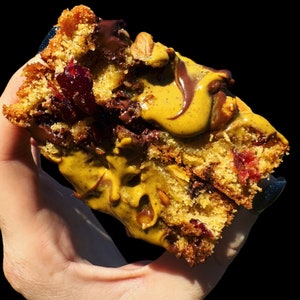 May include: A close-up of a cookie with chocolate chips, cranberries, and nuts. The cookie is topped with a layer of peanut butter and drizzled with chocolate.