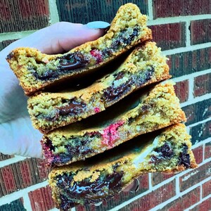 Puede incluir: Una pila de cuatro galletas de chocolate con chips de chocolate, mermelada de frambuesa y relleno de chocolate. Las galletas son de color marrón dorado y tienen una textura crujiente.