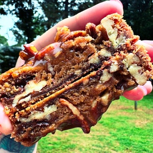 Puede incluir: Primer plano de un brownie de nueces pecanas. El postre está hecho con capas de chocolate y cubierto con nueces pecanas y un chorrito de caramelo. El brownie es de color marrón intenso y parece recién horneado.